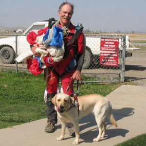 John in his flight suit holding his parachute with guide dog, Tia, by his side